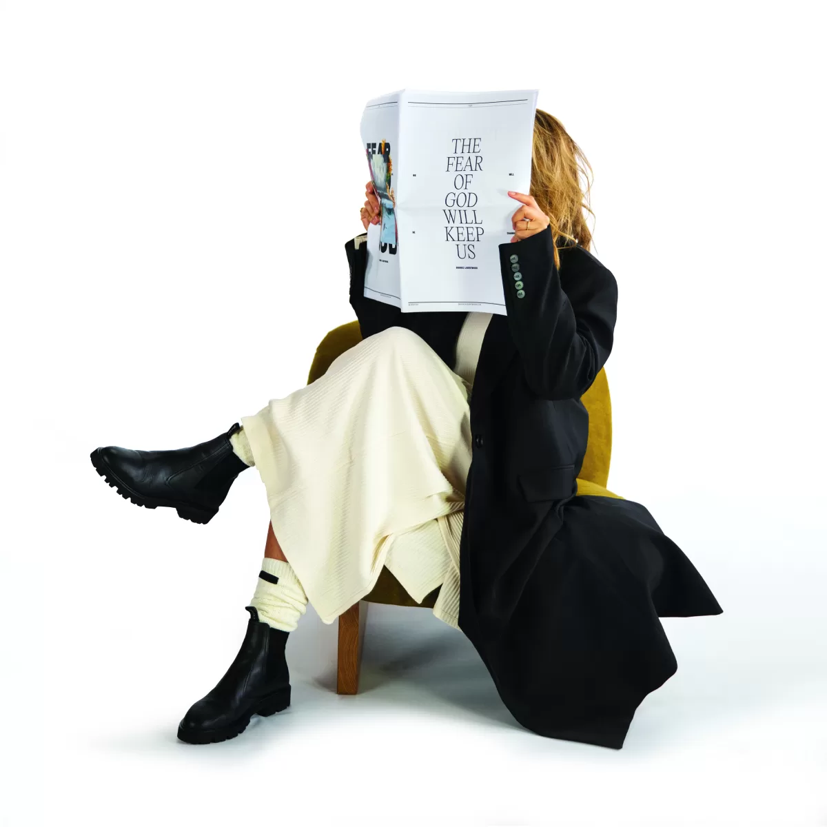 An image of musician and singer, Brooke Ligertwood, sitting down with her legs crossed, and holding a magazine or newspaper in front of her face