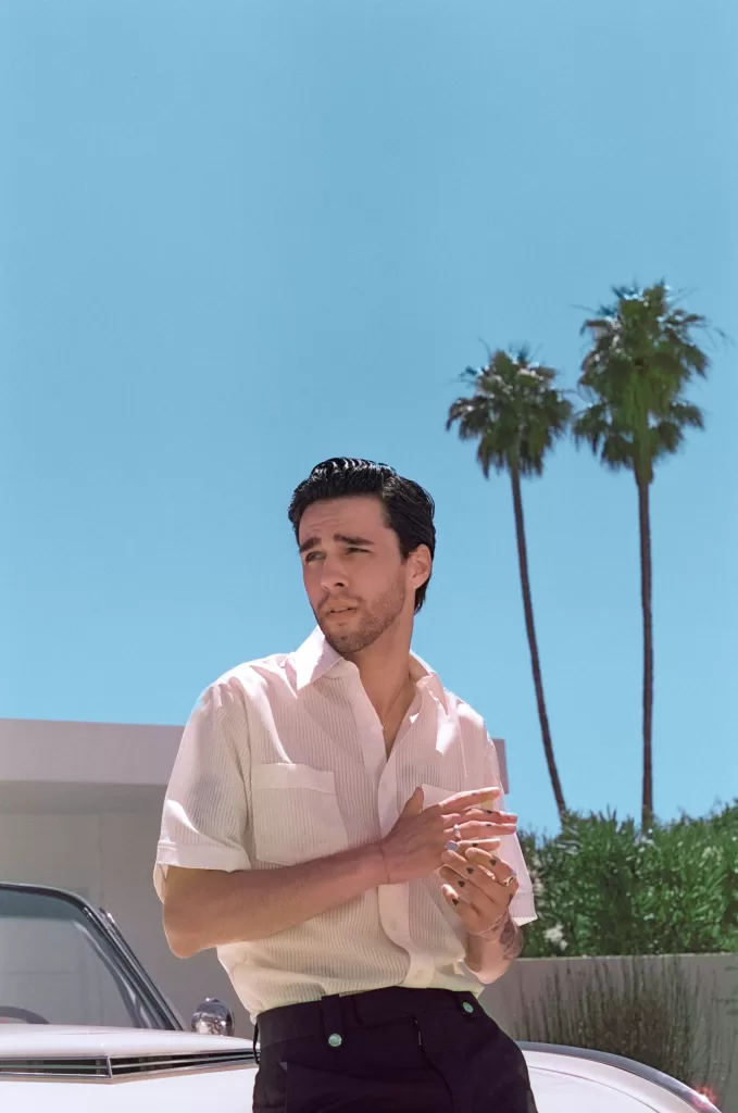 An image of musician and singer, Stephen Sanchez, leaning on hood of car, with palm trees in the background