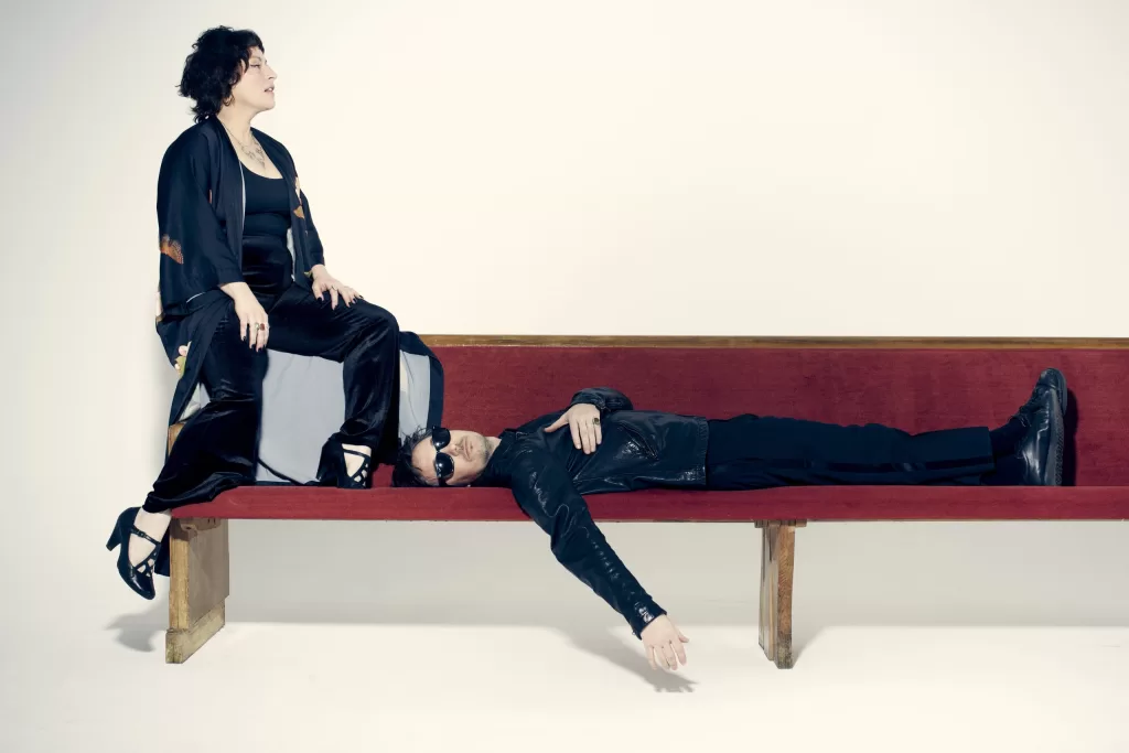 An image of musicians, Cary Ann Hearst and Michael Trent of Shovels and Rope, with Michael laying on a bench, and Cary sitting on top of the edge of the bench