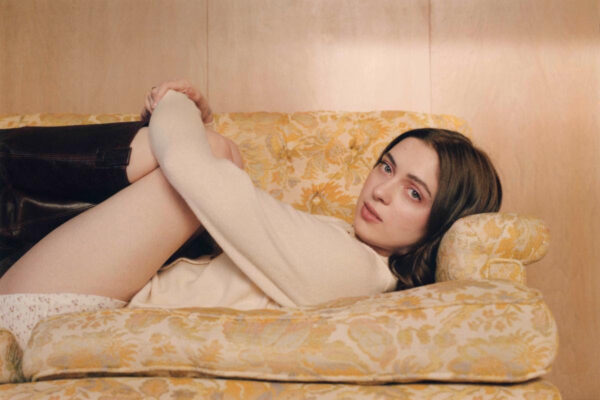 An image of singer-songwriter, Lizzy McAlpine, laying on one side of a sofa end, with her knees up and looking at the camera