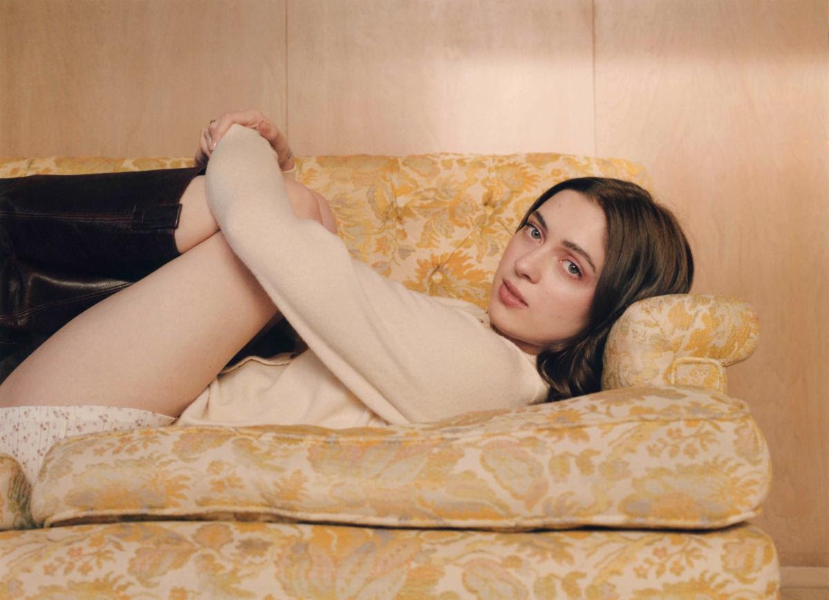 An image of singer-songwriter, Lizzy McAlpine, laying on one side of a sofa end, with her knees up and looking at the camera